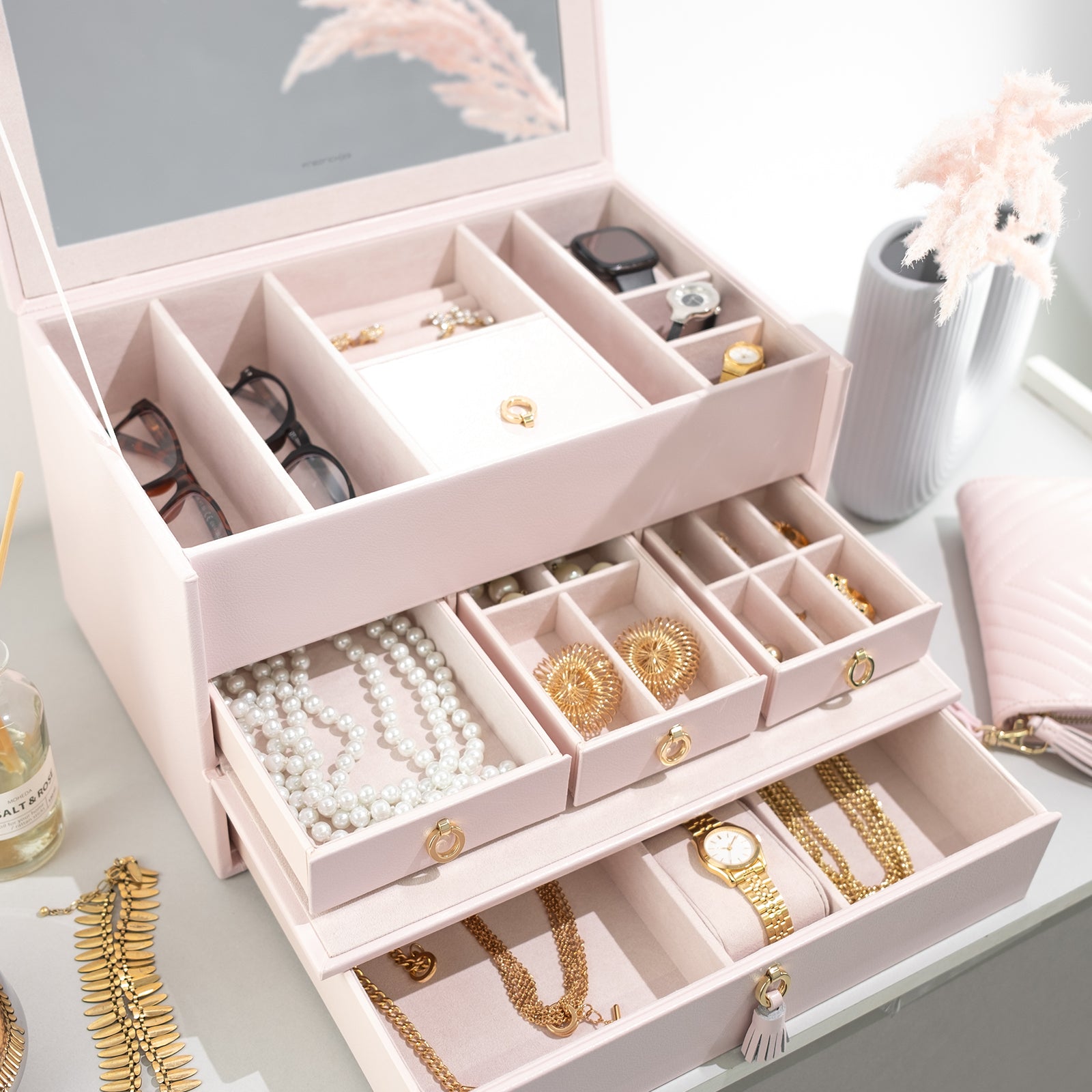 Geöffneter rosa Leder Schmuckkoffer mit Schmuck, Uhren, Brillen und Smartwatch, dekoriert mit Wohnaccessoires.