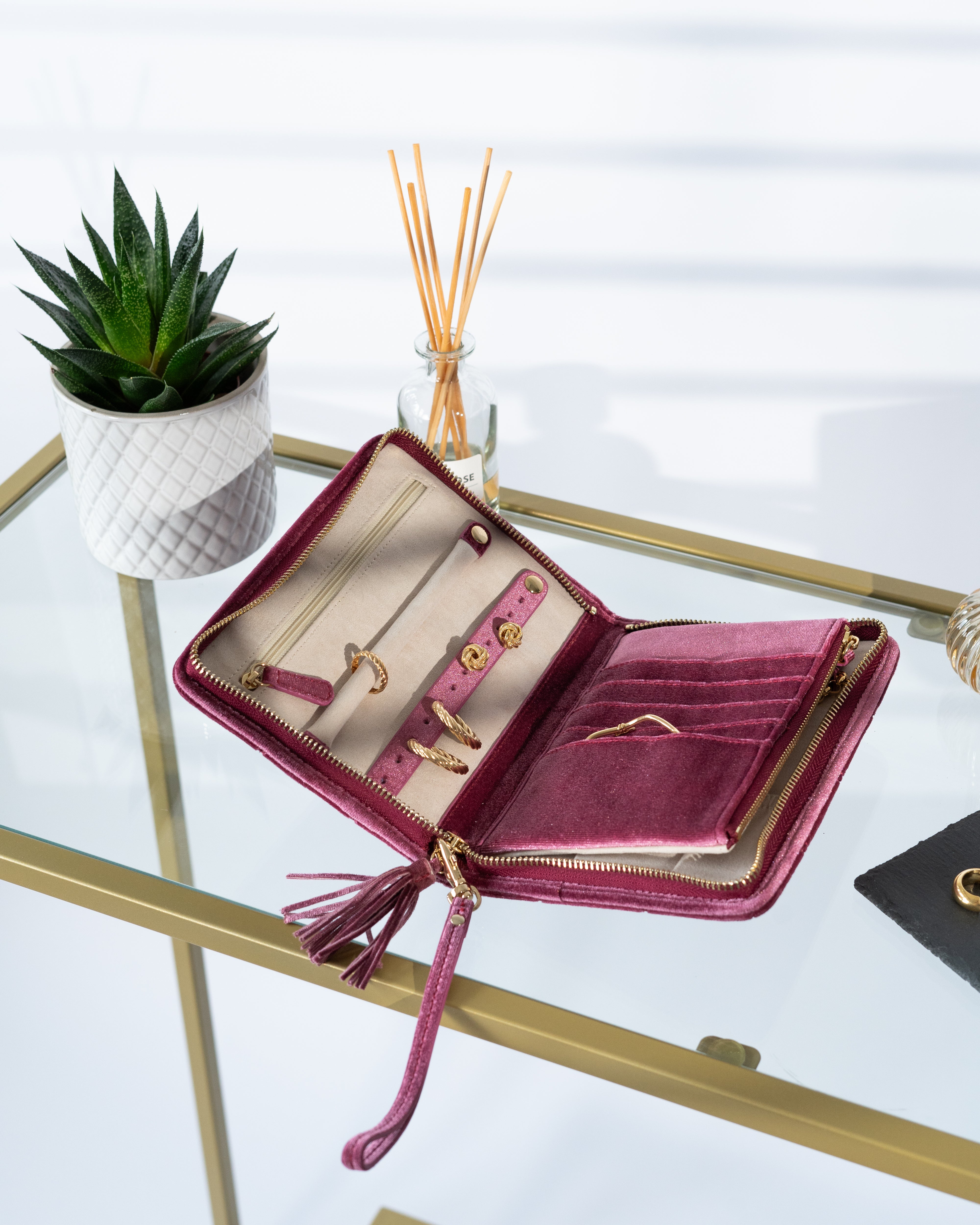 Schmucketui aus Samt in Fuchsia mit Steppmuster, Zierquaste und Handschlaufe auf Glastisch mit Schmuck befüllt.