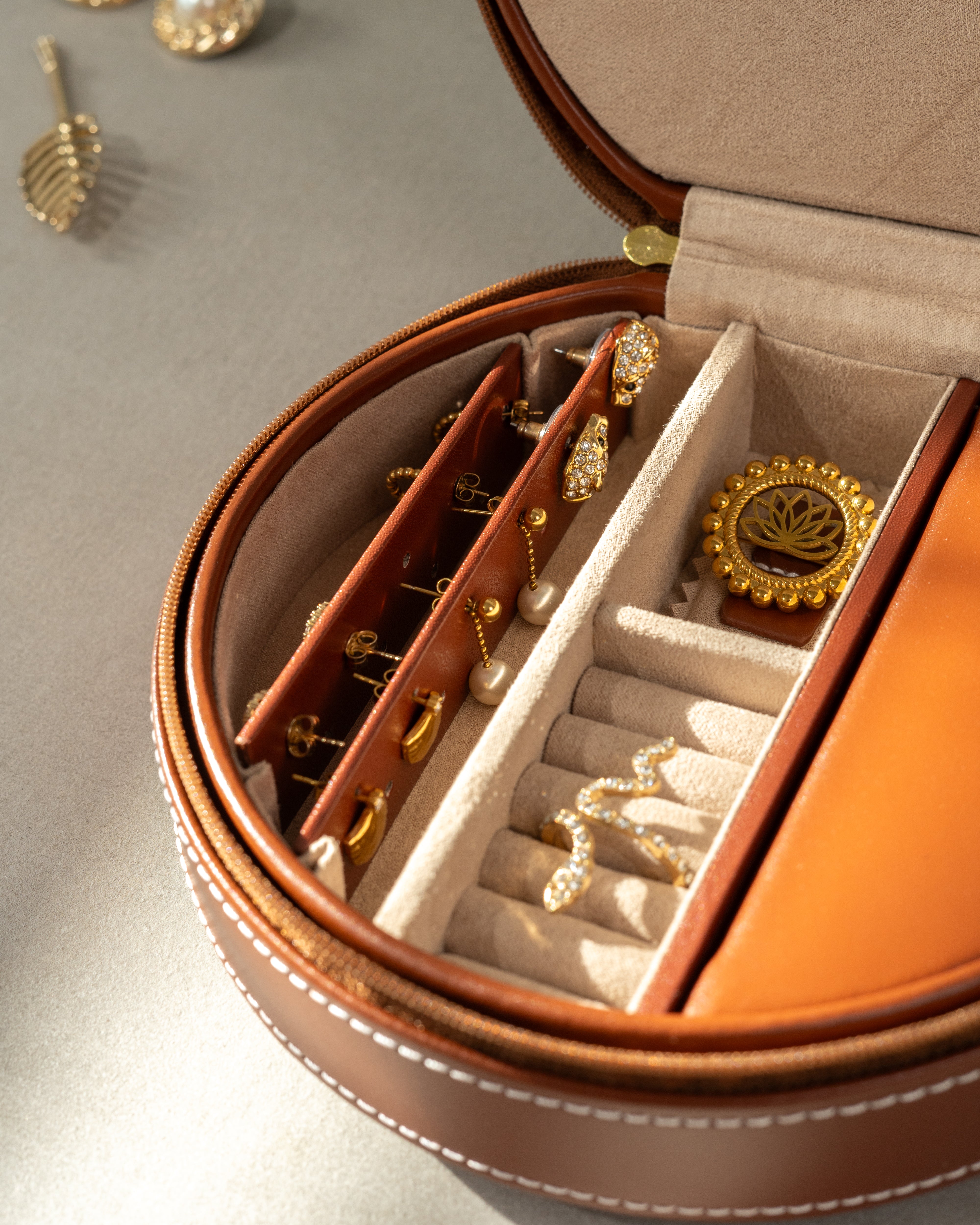 Geöffnetes, braunes, rundes Schmucketui aus Leder mit weißen Ziernähten und Schmuck befüllt.