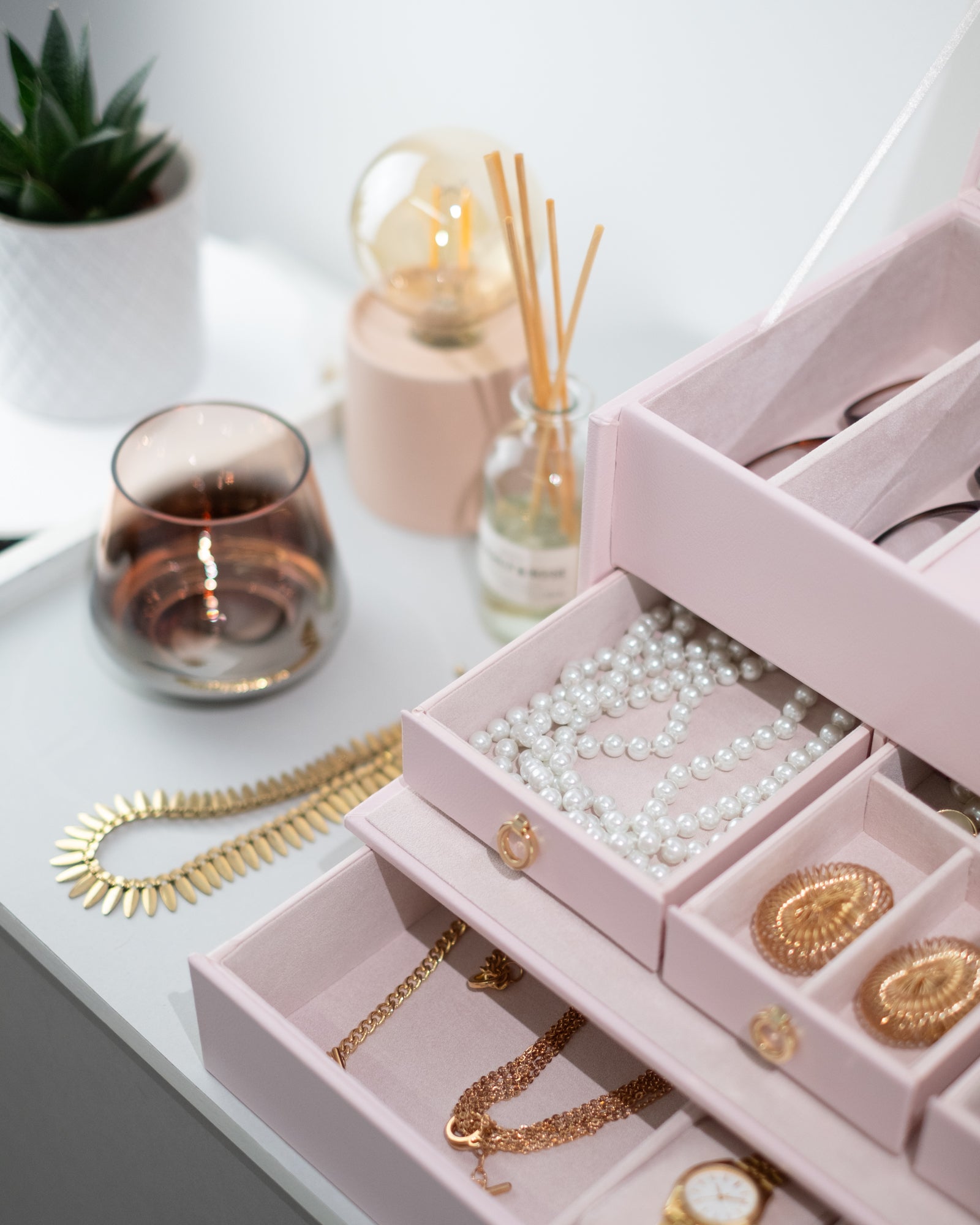 Detailaufnahme von geöffnetem rosa Schmuckkoffer aus Leder befüllt mit Schmuck, Uhren und Brillen. Accessoires im Hintergrund.