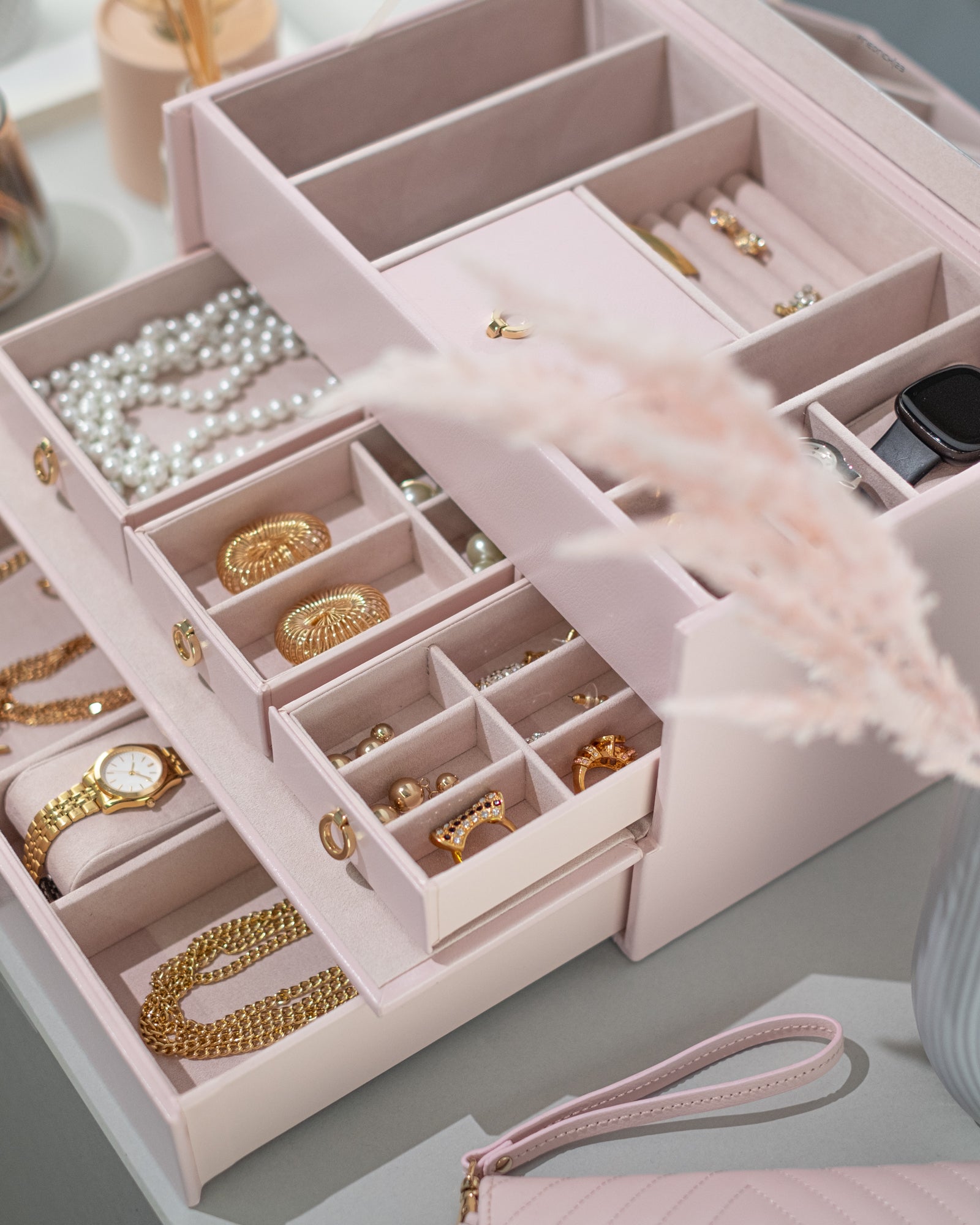 Detailaufnahme von geöffnetem rosa Schmuckkoffer aus Leder befüllt mit Schmuck, Uhr, Smartwatch und Brillen. Accessoires und Schmucketui daneben.