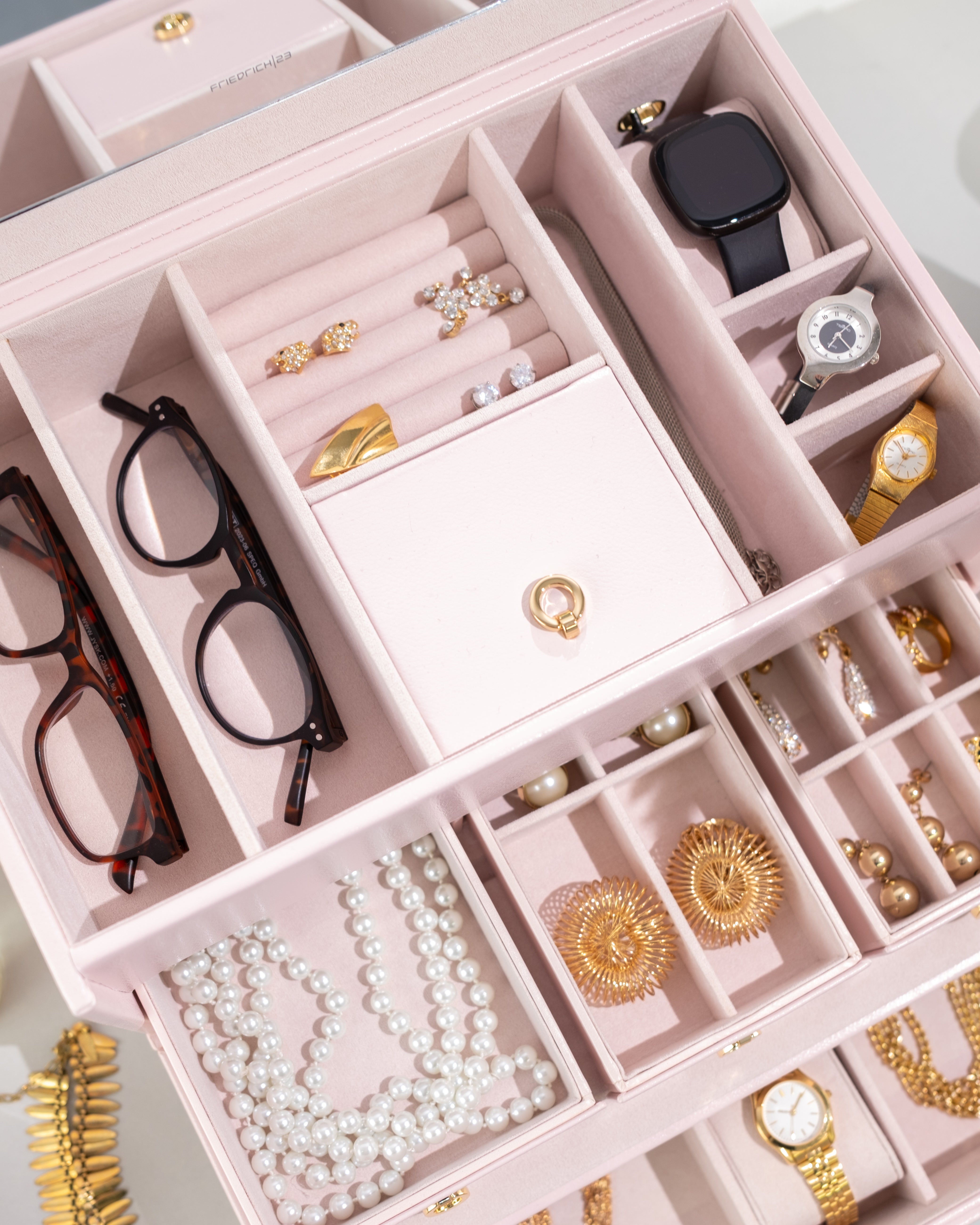 Detailaufnahme von oben von geöffnetem rosa Schmuckkoffer aus Leder befüllt mit Schmuck, Uhr, Smartwatch und Brillen.