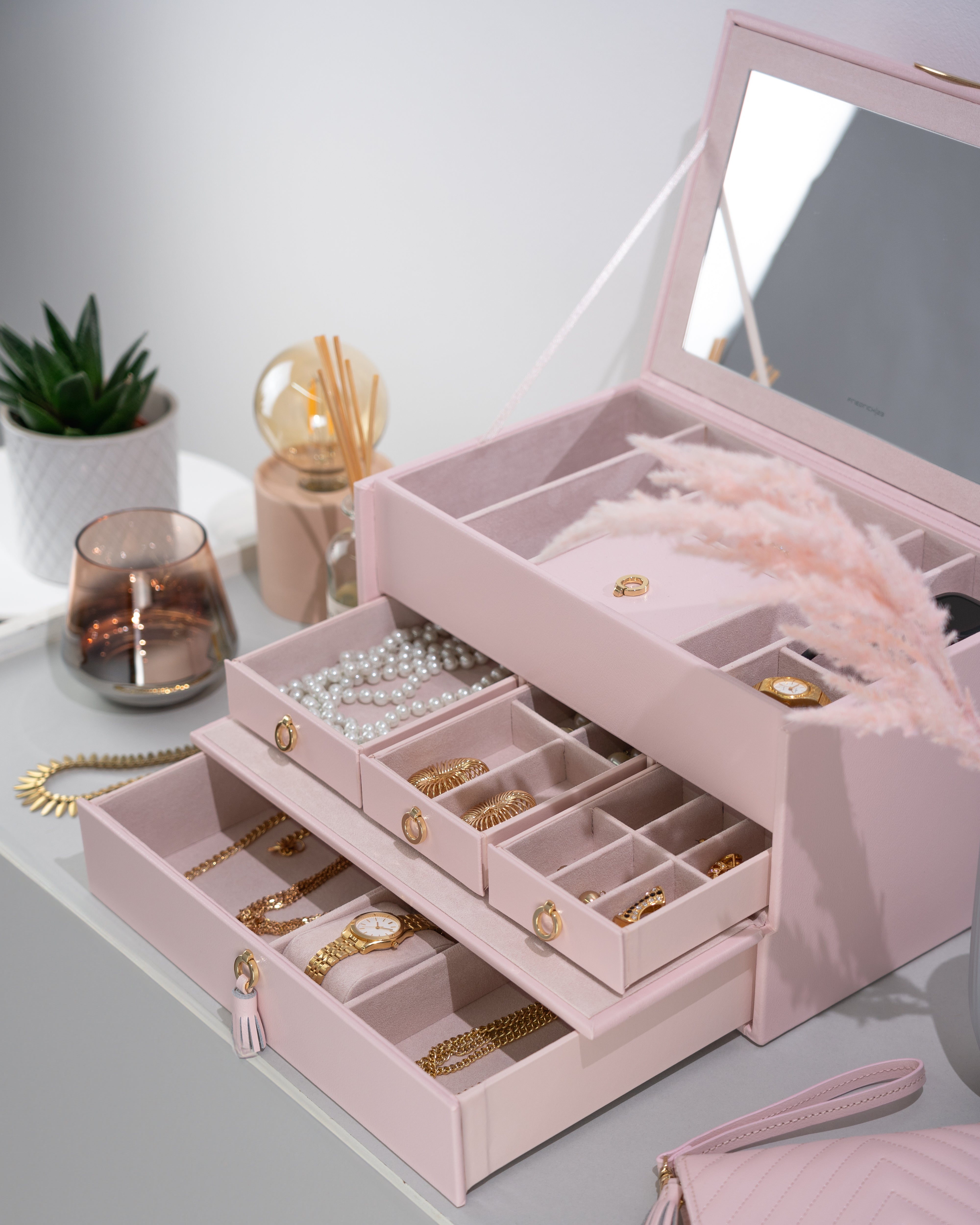 Rosafarbener Schmuckkoffer aus Leder mit Ziernähten und Beschlägen in Goldoptik, Lade mit Zierquaste sowie Friedrich23 Logo in Goldprägung auf Tisch neben Accessoires und Schmucketui.