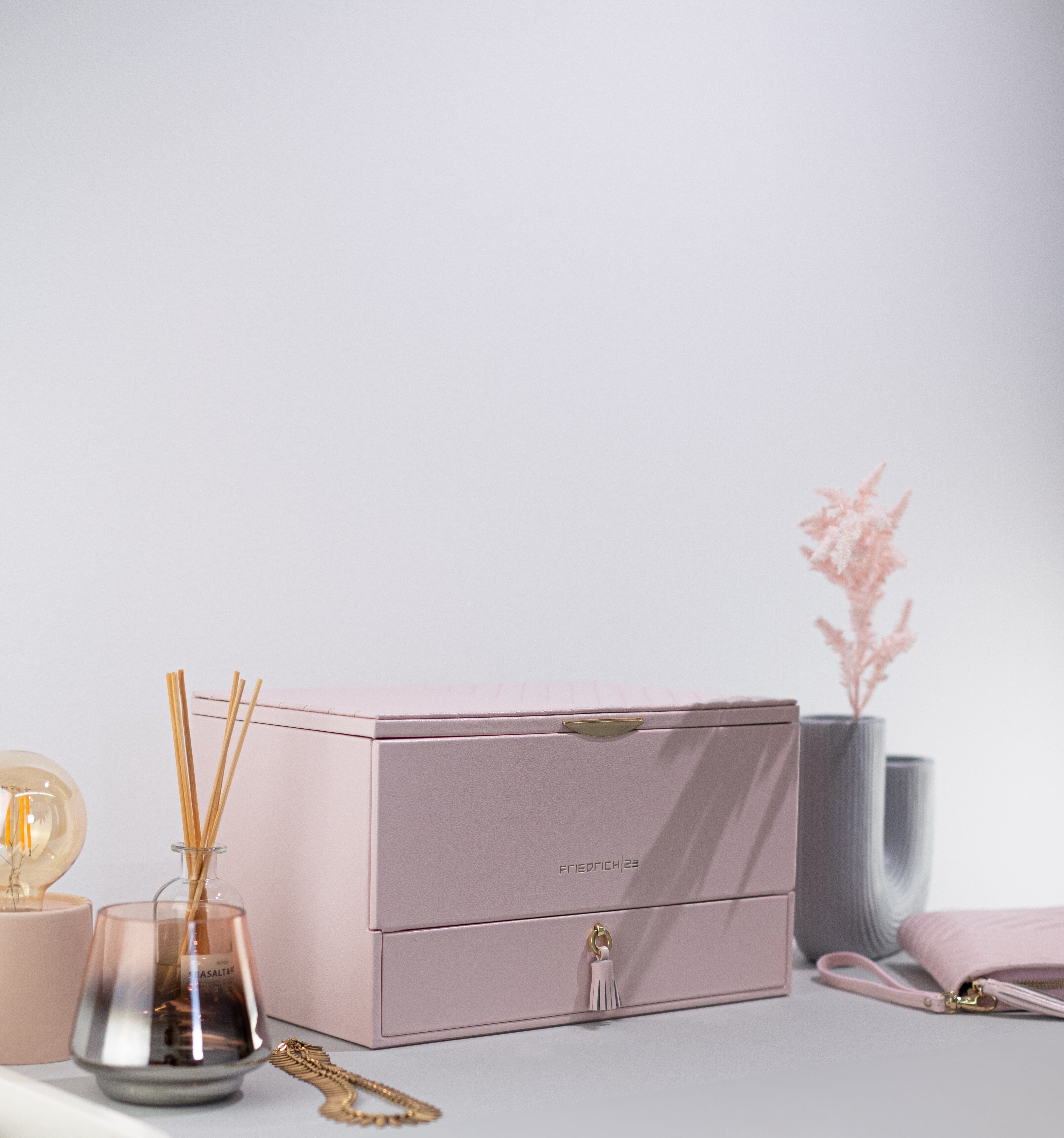 Rosafarbener Schmuckkoffer aus Leder mit Ziernähten und Beschlägen in Goldoptik, Lade mit Zierquaste sowie Friedrich23 Logo in Goldprägung auf Tisch neben Accessoires und Schmucketui.