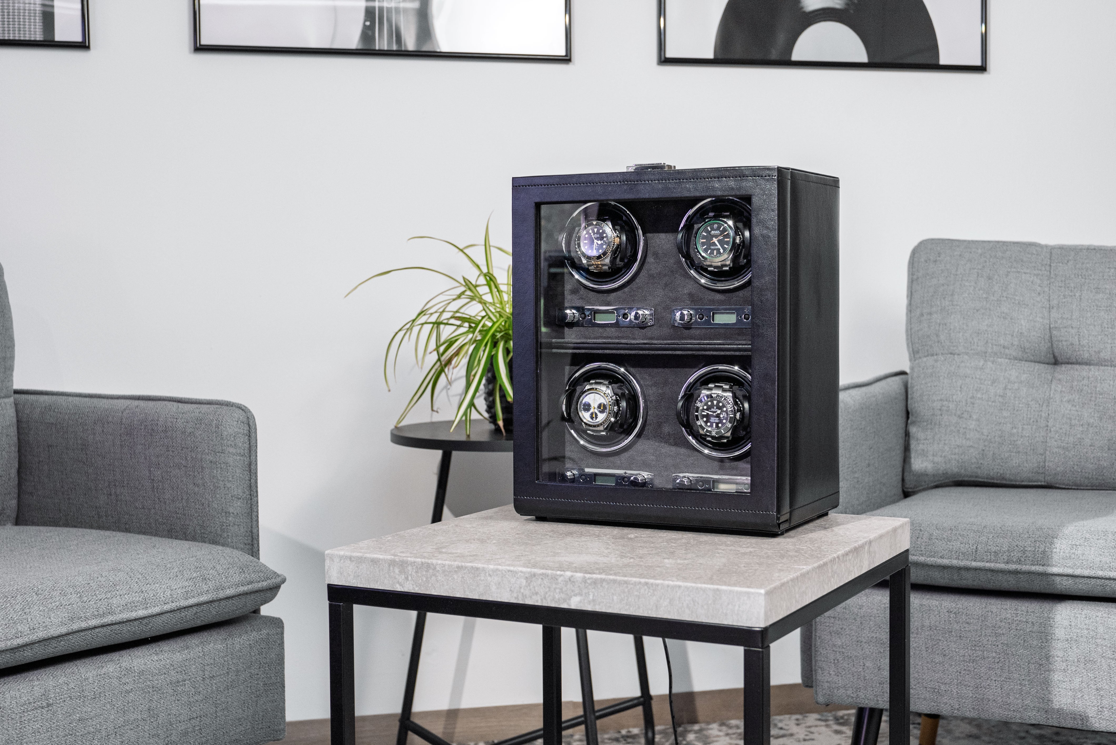 Uhrenbeweger aus schwarzem Leder mit Glasfenster für 4 Uhren, schräg auf Tisch stehend. Wohnsituation mit Sofa bzw. Sessel, Bildern und Pflanze.