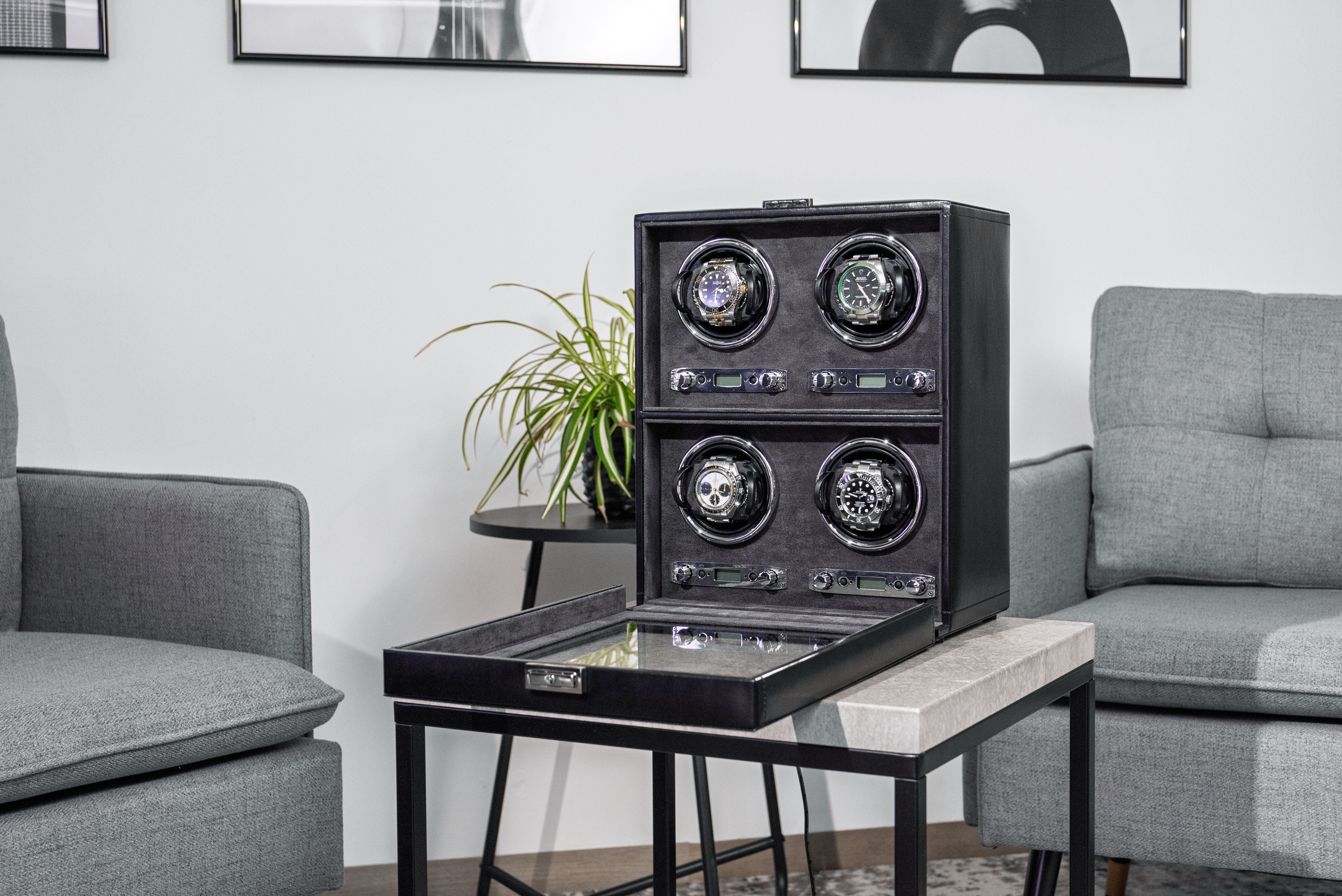 Uhrenbeweger aus schwarzem Leder mit geöffnetem Glasfenster für 4 Uhren, schräg auf Tisch stehend. Wohnsituation mit Sofa bzw. Sessel, Bildern und Pflanze.
