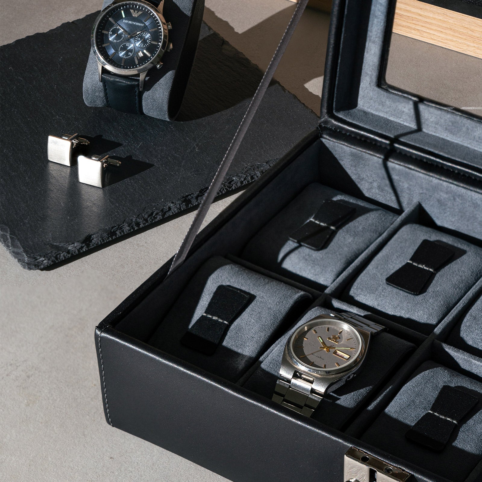 Geöffnete Leder Uhrenbox mit Uhren, Uhrenkissen und männlichen Accessoires daneben.