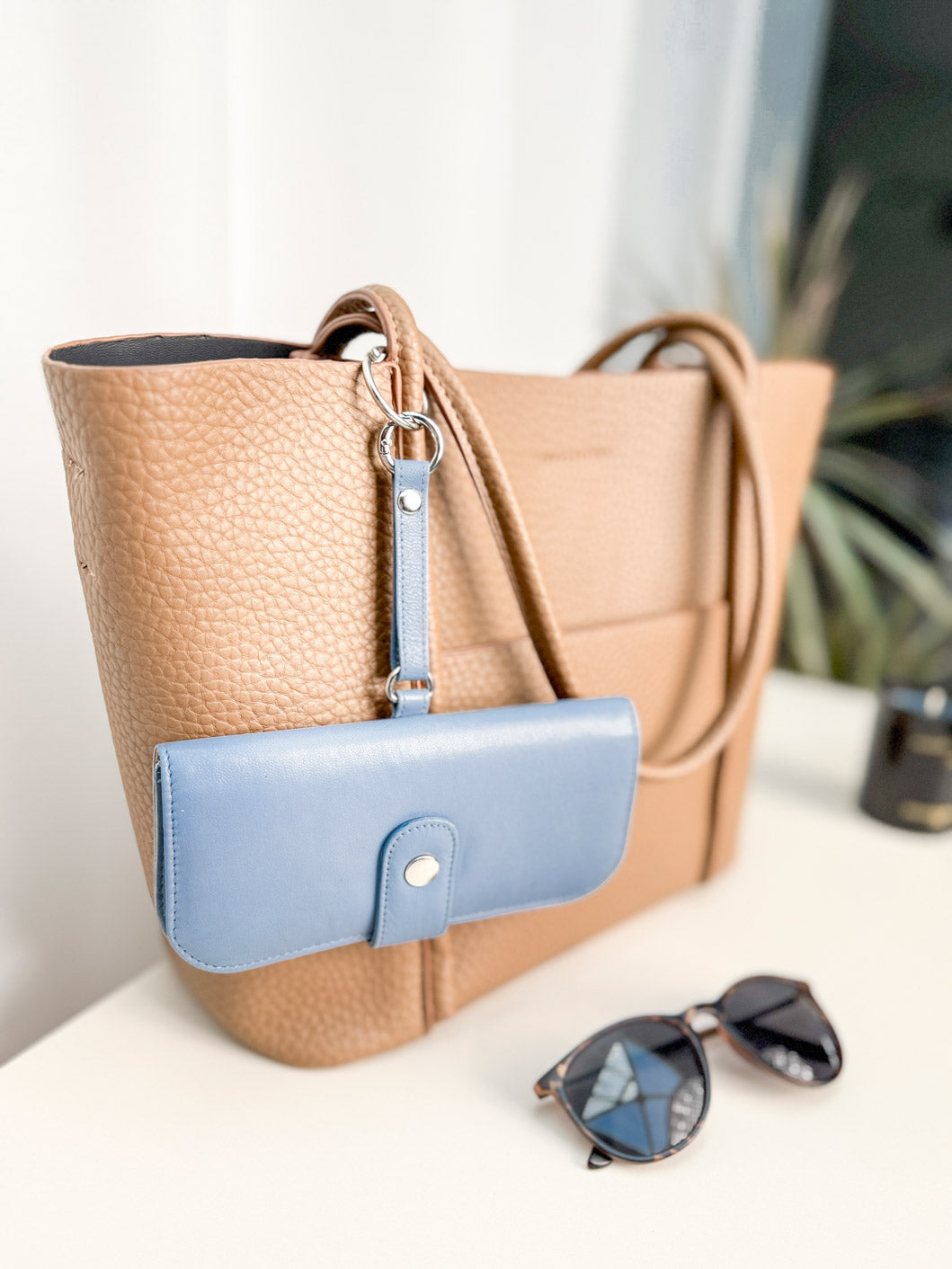 Blaues Brillenetui hängt an einer Handtasche mit einem kleinen runden Karabiner befestigt. Daneben liegt eine Sonnenbrille.