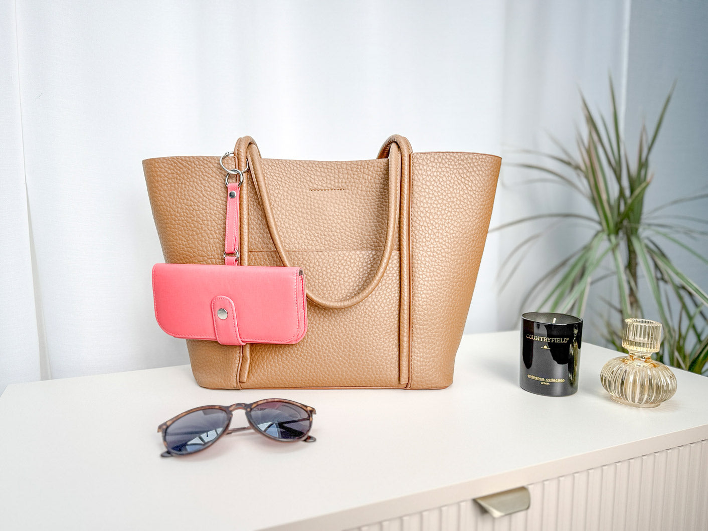 Brillenetui hängt an einer Handtasche mit einem kleinen runden Karabiner befestigt. Daneben liegt eine Sonnenbrille. Auf der Kommode steht eine Kerze sowie ein Parfüm.