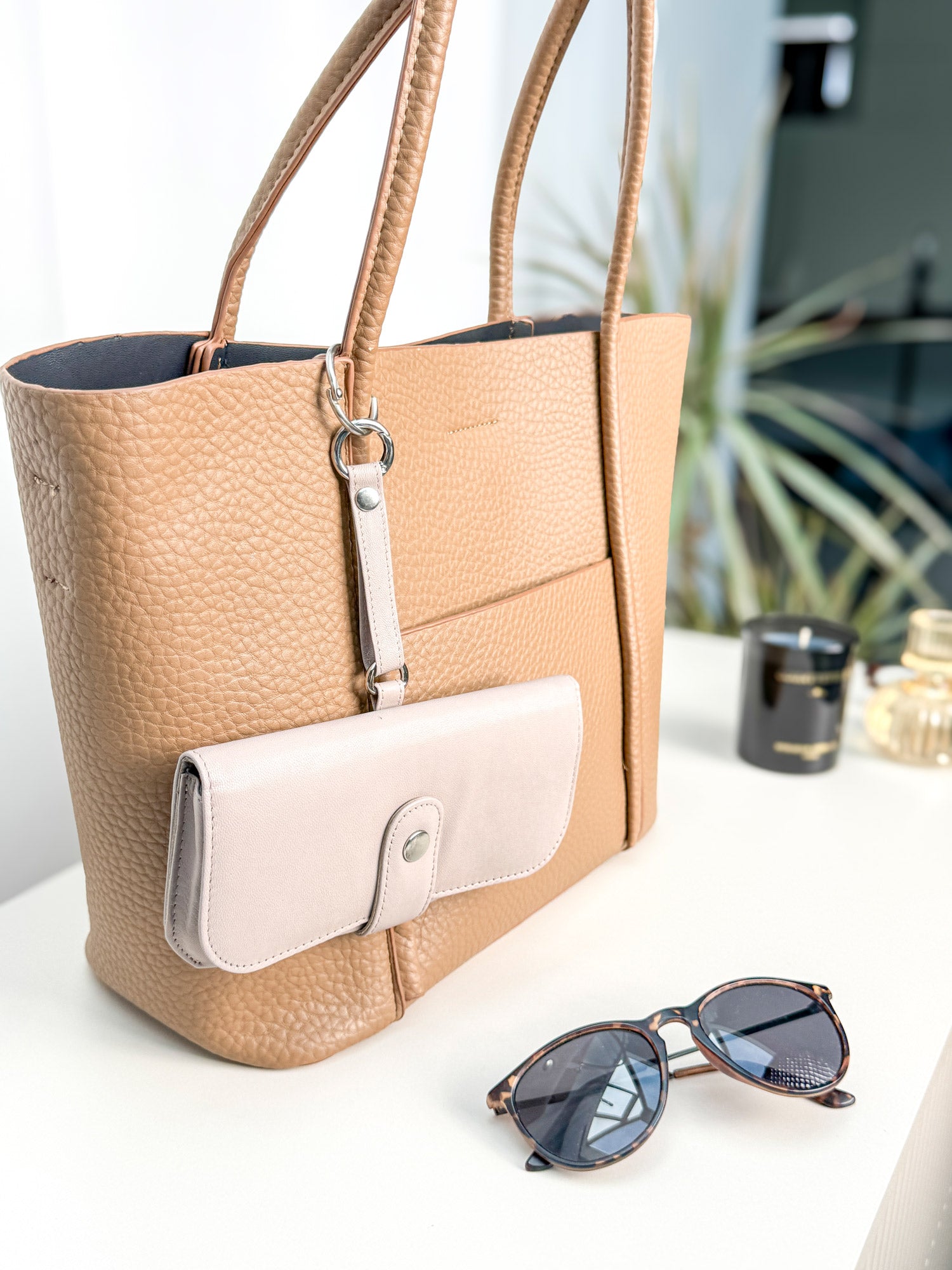 Beiges Brillenetui hängt an einer Handtasche mit einem kleinen runden Karabiner befestigt. Daneben liegt eine Sonnenbrille.