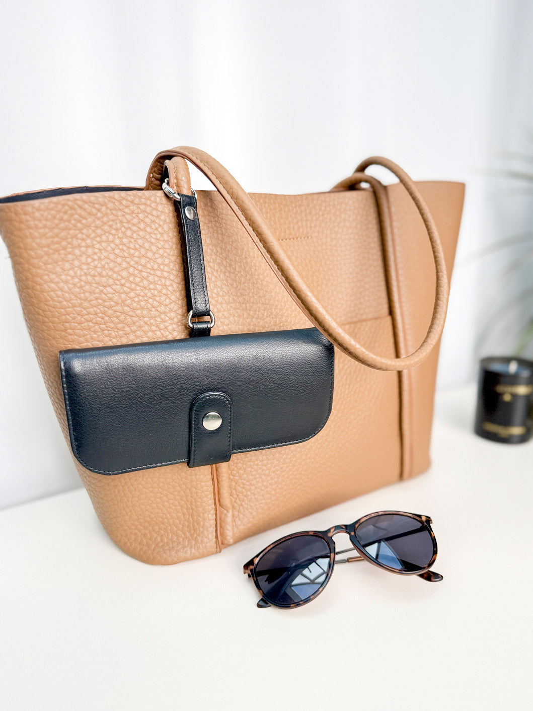 Schwarzes Brillenetui hängt an einer Handtasche mit einem kleinen runden Karabiner befestigt. Daneben liegt eine Sonnenbrille.