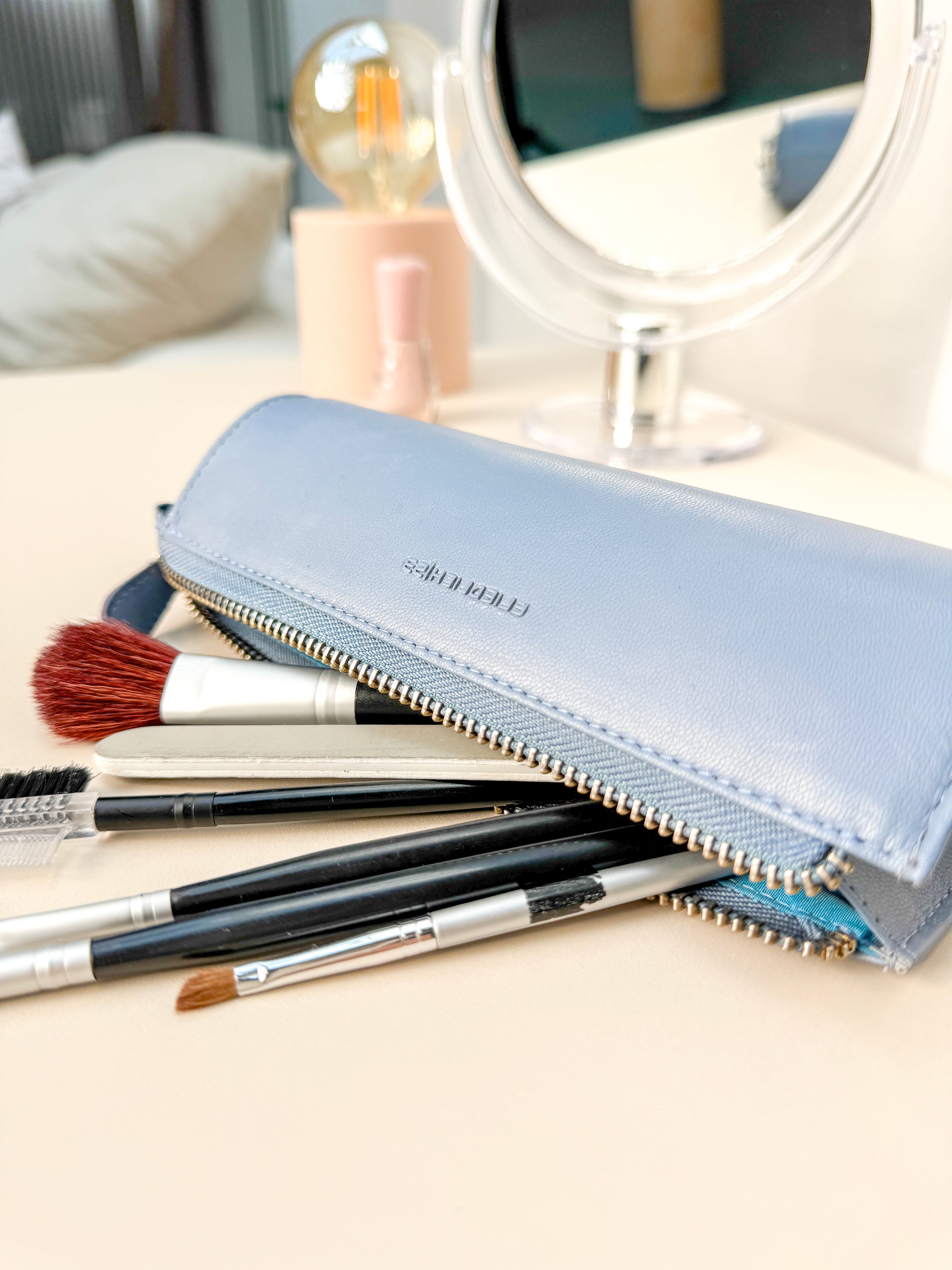 Beauty-Stifterolle aus Leder liegt geöffnet auf einer Kommode mit Spiegel und Beautyartikeln, der Reißverschluss ist geöffnet und mehrere Schminkpinsel sind sichtbar.